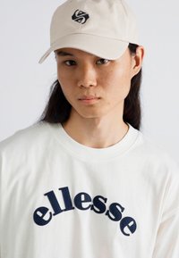 Camiseta blanca con logo de "ellesse" en navy curvado, combinada con una gorra beige que presenta un logo negro. La tela es suave y el diseño es informal.
