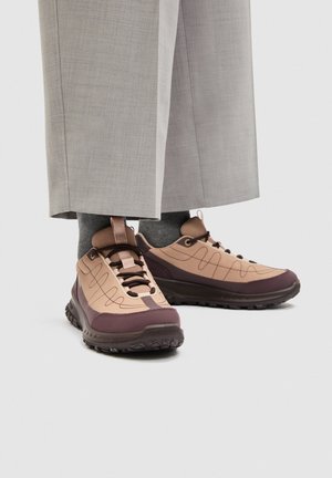 Füße, die beige und dunkelbraune Sneakers mit dunkelgrauen Socken und hellgrauen, weit geschnittenen Hosen auf weißem Hintergrund tragen.