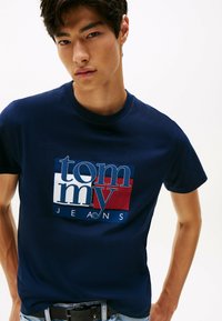 Joven vistiendo una camiseta Tommy Jeans azul marino con logo, combinada con un vaquero azul claro y un cinturón negro, contra un fondo blanco.