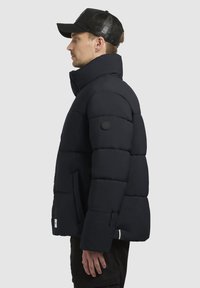 Tumesin puffer-jakk, millel on kõrge krae, tikanditekstuur, küljetaskud ning logotäht küljepeal. Kandsin musta võrkkorki.