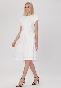 Robe en coton blanche avec une jupe à volants, des manches courtes, un col rond et un subtil détail de couture au bas. Portée avec des escarpins noirs.