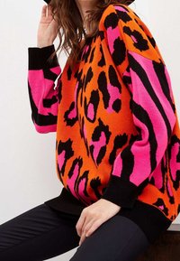 Kobieta ubrana w sweter w jasno pomarańczowy i różowy wzór w leopardzie z czarnymi mankietami i dołem, w połączeniu z czarnymi spodniami, siedząca na drewnianej powierzchni.