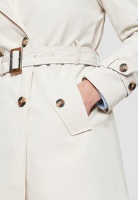 Beige trenčkot s páskem v pase, předním zapínáním na knoflíky a bočními kapsami. Obsahuje zlaté knoflíky a hladkou strukturu tkaniny.