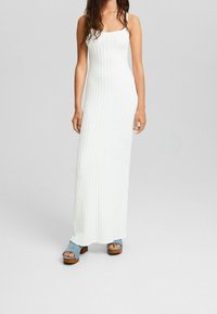 Robe maxi longue blanche côtelée avec fines bretelles. Détails remarquables : motif texturé et silhouette ajustée. Assortie à des sandales épaisses à bout ouvert.