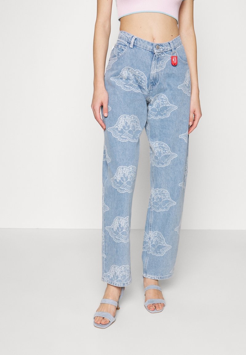 Femme portant un jean taille haute bleu clair avec des motifs d'anges blancs, un crop top rose et des sandales à talons bleu clair.