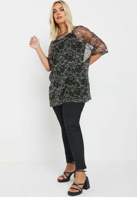 Top in pizzo nero con maniche a tre quarti, caratterizzato da motivi floreali e una vestibilità ampia, abbinato a pantaloni neri slim e sandali neri chunky.