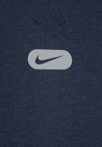 La sudadera azul marino presenta un tejido texturizado con un logotipo de Nike de color gris claro y suave en forma ovalada, exhibido de manera centrada en la parte frontal.