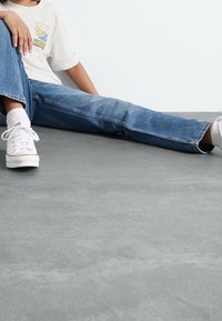 Vybledlé modré džíny rovného střihu, doplněné bílými teniskami, nošeny s světle barevným tričkem s potiskem. Nohy jsou překřížené na šedém povrchu.