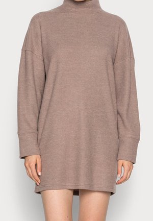 Frau trägt ein lockeres, langärmliges, hellbraunes Rollkragenpulloverkleid mit geradem Saum über den Knien.