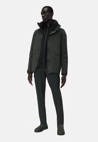 Giacca impermeabile verde scuro con colletto alto, chiusura con zip e bottoni a pressione. Abbinata a pantaloni aderenti e stivali alla caviglia.