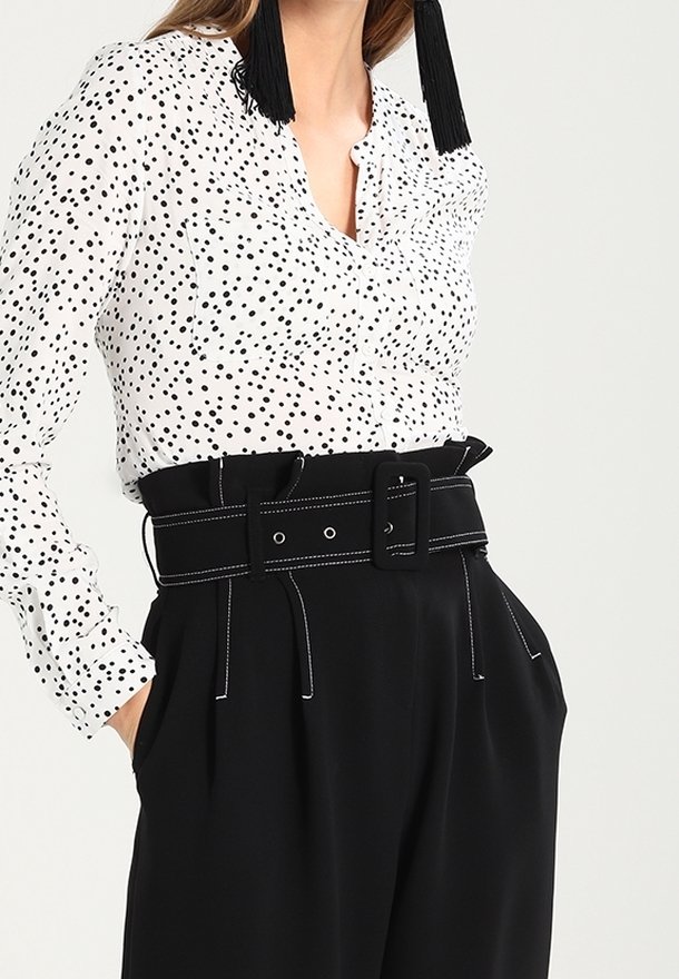 Femme portant une blouse blanche à manches longues avec des pois noirs, un pantalon noir taille haute avec une large ceinture et des boucles d'oreilles noires à pompons.