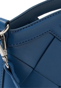 Niebieska skórzana torebka o geometrycznym wzorze, gładkiej teksturze i metalowych elementach, z widocznym przeszyciem i wyraźnie zdefiniowanym kształtem.