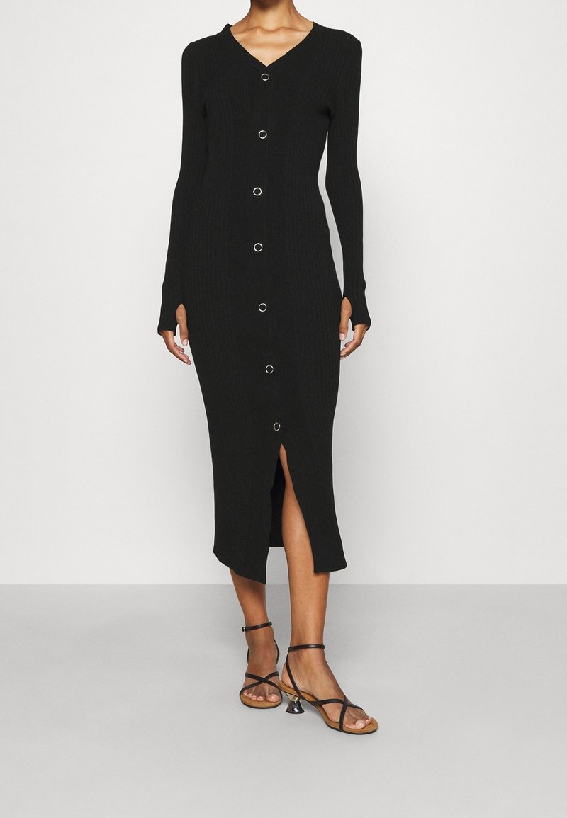Abito midi in maglia nera a coste con scollatura a V, maniche lunghe, bottoni frontali con hardware circolare e spacco laterale, abbinato a sandali neri con cinturini.