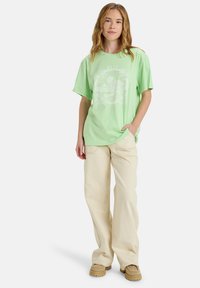 Hellgrünes Oversize-T-Shirt mit einem weißen Berggrafik, kombiniert mit lockeren cremefarbenen Hosen und beigen Slippern.
