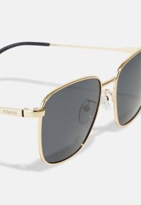 Ochelari de soare Polaroid cu un cadru metalic auriu, accente negre și lentile gri închis. Caracteristicile includ pernuțe de nas ajustabile și un design elegant.