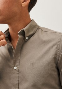 Helles braunes Hemd mit Knöpfen und subtiler Textur, mit einem Kragen und einem gestickten Logo auf der Brust. Eierschalenfarbene Knöpfe verleihen dem Hemd das besondere Etwas.