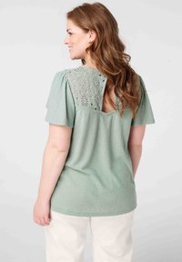 Mintgroene top met textuurstof, korte pofmouwen en een kanten paneel op de rug met cirkelvormige patronen.