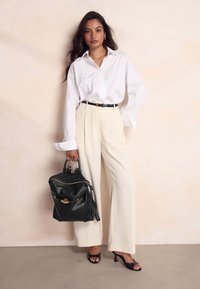 Witte blouse, crèmekleurige wijde broek, zwarte riem en zwarte leren rugzak met gouden accenten. Model op hakken met lang haar.