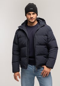 Granatowa kurtka puchowa z zamkiem z przodu, kapturem i pikowanym wzorem. Nosi się ją na granatowym swetrze. Połączona z jasnoniebieskimi dżinsami i czarną czapką beanie.