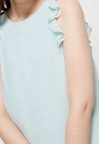 Abito senza maniche azzurro chiaro con dettagli arricciati sulle spalle. Tessuto liscio e silhouette dritta, che mette in risalto la spalla e il braccio nudi.