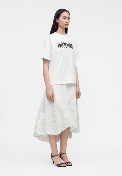Γυναίκα που φοράει λευκό t-shirt Moschino, λευκή ασύμμετρη φούστα με ρίγες και μαύρα σανδάλια με τακούνι, στέκεται μπροστά σε απλό φόντο.