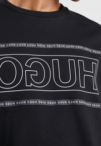 Schwarzes Baumwoll-T-Shirt mit einem auffälligen weißen Logodesign mit wiederholtem Text, Rundhalsausschnitt und kurzen Ärmeln.
