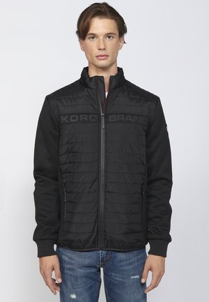 Veste noire avec texture matelassée sur le devant, manches tissées et col montant. Comprend des zippers et le logo de la marque sur la poitrine. Un jean en denim est visible.