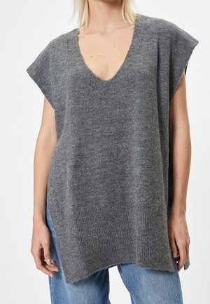 Kobieta ubrana w luźny szary, dziergany bezrękawnik z głębokim dekoltem w kształcie litery V i niebieskie dżinsy na jednolitym tle.