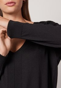 Czarny, monochromatyczny sweter z dekoltem w serek, o fakturowanej tkaninie, z prążkowanymi mankietami i długimi rękawami, częściowo zakryty przez dłoń.