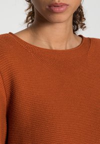 Rust-colored sweater with a textured knit pattern and a wide round neckline, displaying soft, lightweight material.