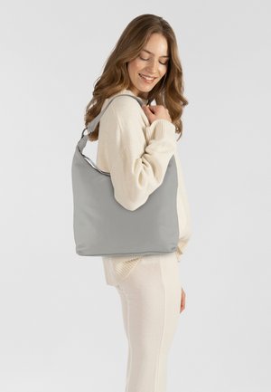 Graue Schultertasche aus glattem Material, ausgestattet mit einem verstellbaren Trageriemen und einem minimalistischen Design, gehalten von einer Person in einem cremigen Pullover.