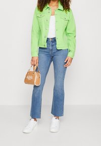 Neongrön jeansjacka med silverknappar, vit t-shirt, utsvängda blå jeans, vita sneakers och en beige handväska med logotyp.