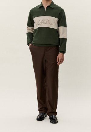 Mann trägt dunkelgrünes und beiges Langarm-Poloshirt mit dem Text "Les Deux", braune Hose und schwarze Schuhe, steht mit Händen in den Taschen.