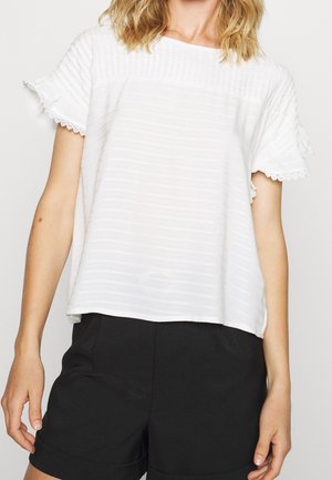 Vrouw die een witte korte mouwen blouse met structuur en kanten rand draagt, samen met zwarte, op maat gemaakte korte broek, tegen een effen achtergrond.