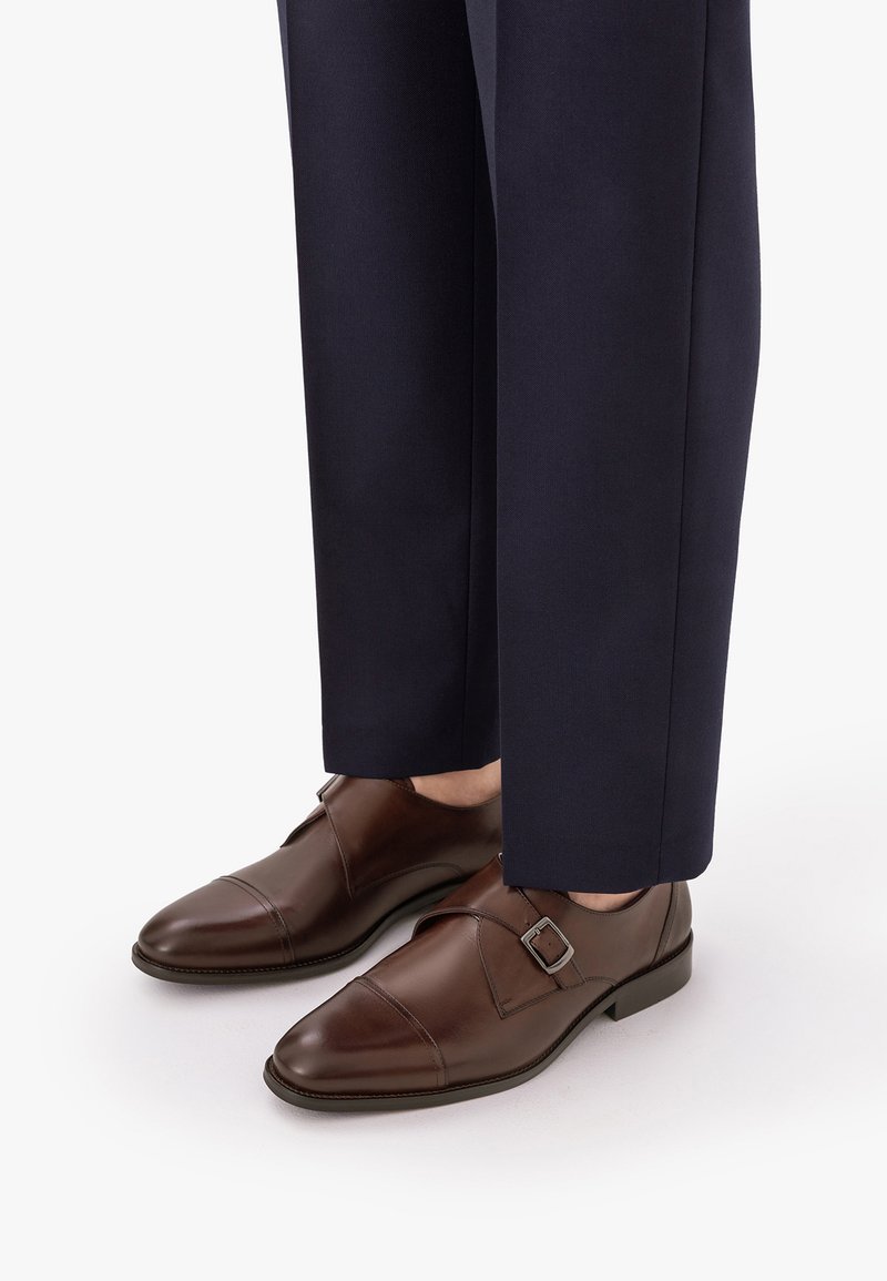 Καφέ δερμάτινα παπούτσια monk strap, φορεμένα με σκούρο μπλε επίσημο παντελόνι σε λευκό φόντο.