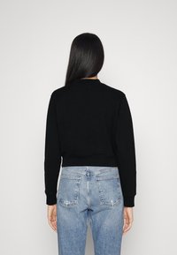 Mujer con cabello largo y oscuro, vistiendo una sudadera corta negra y vaqueros de talle alto azul claro, de pie contra un fondo blanco liso.