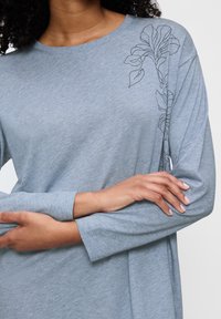 Maglia a maniche lunghe grigia in tessuto morbido con un contorno di disegni floreali sulla spalla sinistra. Scollatura rotonda e vestibilità comoda.