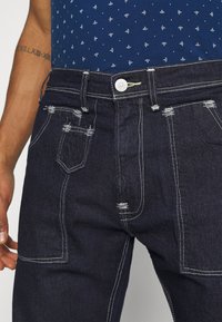 Tummat farkut, joissa on valkoiset ompeleet, kaksi etutaskua, pieni kolikoille tarkoitettu tasku ja pieniä kulumiskohtia taskujen lähellä.