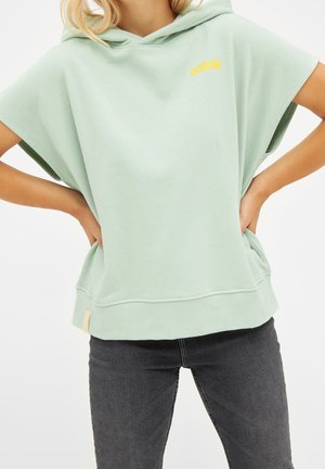 Femme portant un sweat à capuche à manches courtes vert clair avec "DERBE" en jaune sur la poitrine et un jean gris foncé, les mains posées sur les hanches.