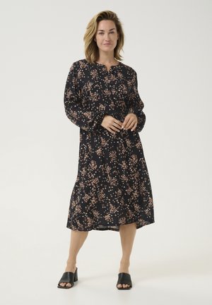 Mustapohjainen kukallinen mekko pitkilla hihoilla, nappikiinnityksellä edessä ja polvimittaisella helmaa; siinä on pieni vaaleanpunainen ja vaaleankerman kukkakuosi.