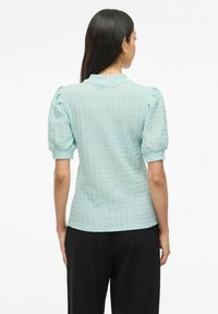 Blusa azzurro chiaro con texture, maniche corte a sbuffo, collo rotondo e silhouette aderente, abbinata a pantaloni neri.