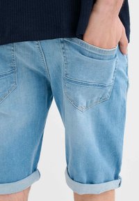Shorts de mezclilla azul claro con dobladillos enrollados, que cuentan con dos bolsillos traseros con un sutil patrón de pespuntes en zigzag. Textura suave y diseño informal.