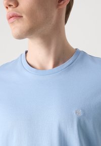 Light blue cotton t-shirt with a crew neckline and short sleeves. Features a small embroidered logo on the chest. Smooth texture.