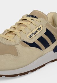 Sneaker Adidas beige e blu navy con dettagli in camoscio e mesh, tre strisce blu navy, lacci beige e logo Adidas sul lato e sulla linguetta.