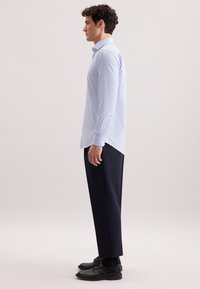 Homme portant une chemise rayée blanche et bleu clair à manches longues, un pantalon sombre et des chaussures de ville noires, se tenant de profil devant un fond uni.