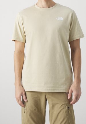 Homme portant un t-shirt à manches courtes beige de The North Face et un pantalon cargo beige avec poches zippées, debout devant un fond uni.