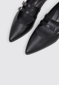 Bailarinas negras de cuero con punta afilada, con dos correas ajustables y hebillas plateadas. Textura suave con un diseño minimalista y sin patrones.