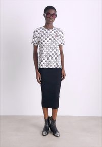 Žena nosi velike sunčane naočale, bijelu majicu s ponavljajućim uzorkom "NY", crnu rebrastu midi suknju i crne gležnjače.