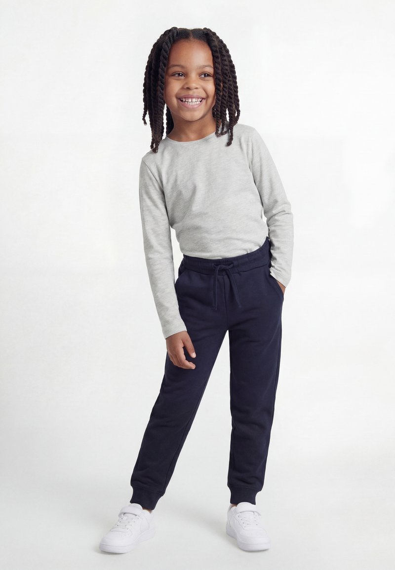 Niño sonriente con cabello trenzado, vestido con una camiseta de manga larga gris, pantalones deportivos azul marino y zapatillas blancas, de pie sobre un fondo blanco.
