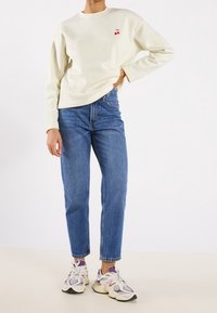 Persona che indossa un felpa color crema con ricamo di ciliegie, jeans blu e sneakers bianche-viola, in piedi con una mano sul fianco.